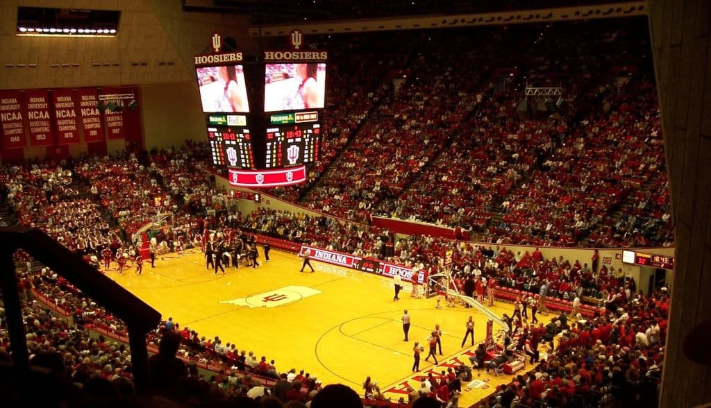 Crowded basketball arena with players on the court, a large scoreboard displaying "Hoosiers," and fans filling the stands. Indiana University banners are visible.