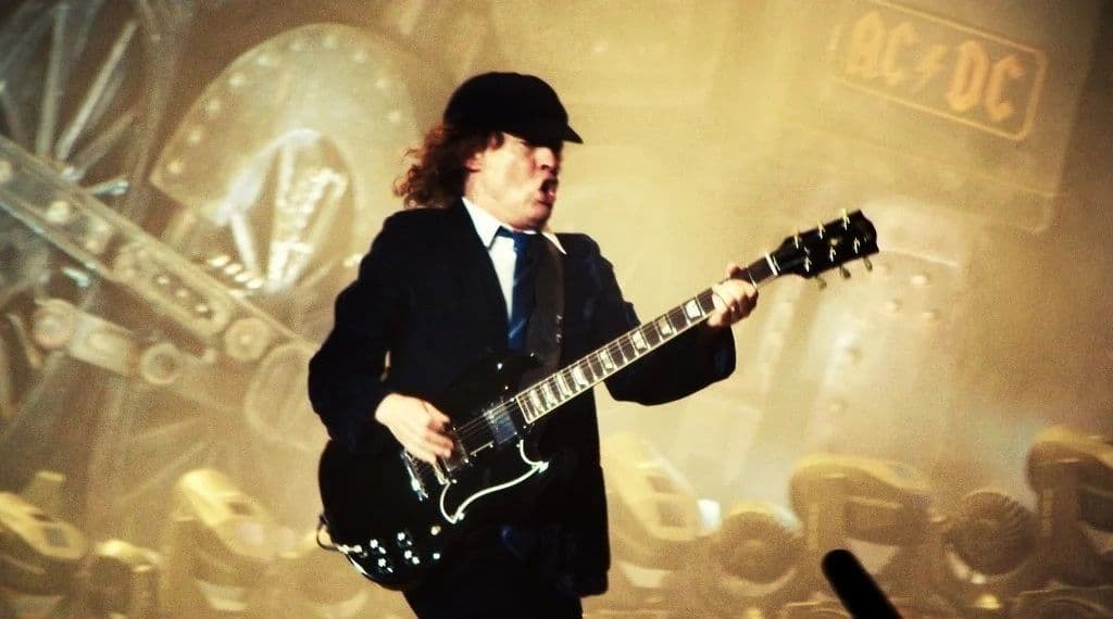 A guitarist in a suit and hat plays an electric guitar on stage with an AC/DC sign and industrial-themed background.